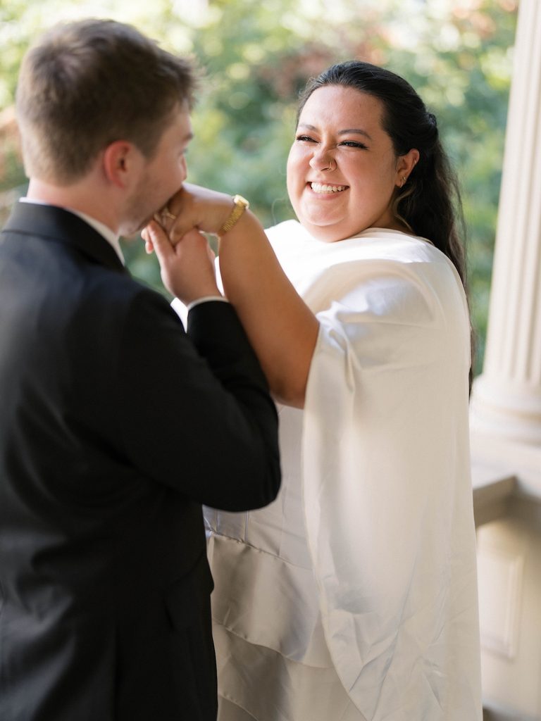 Bride laughing as groom kisses her hand on a porch with classical stone columns in the background.