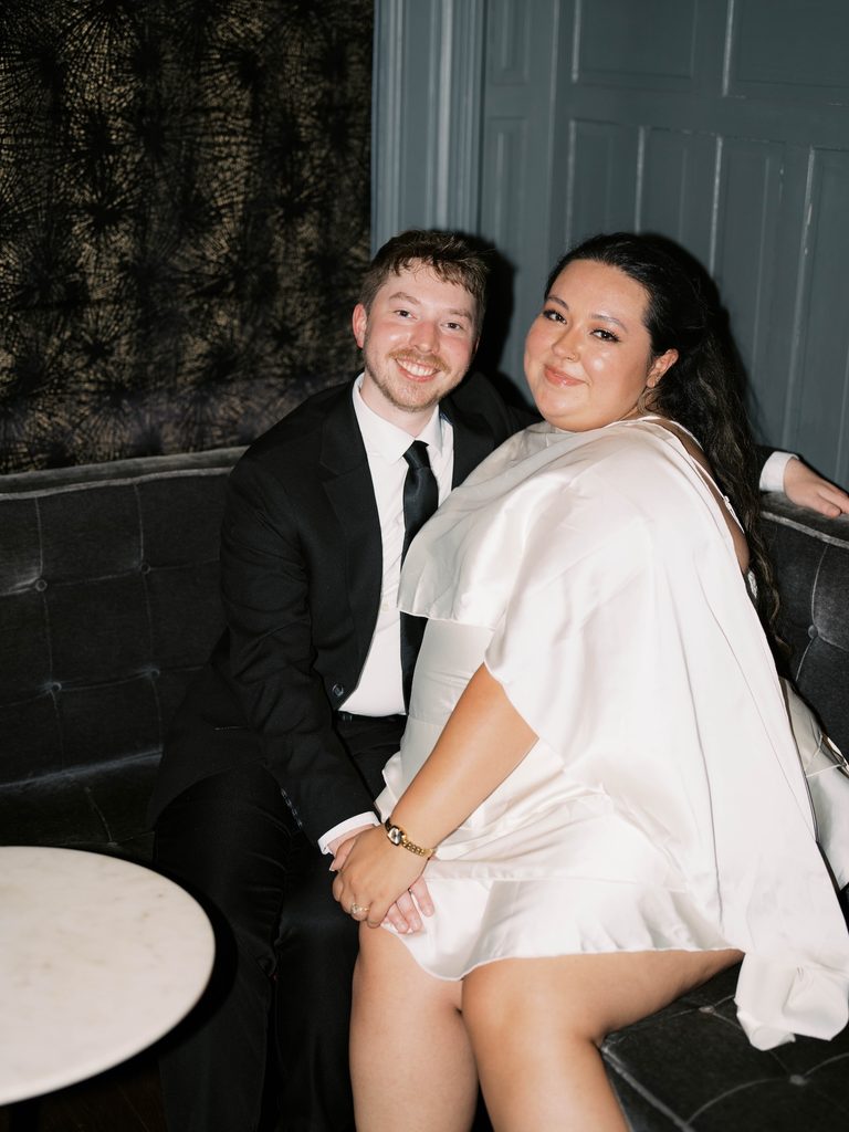 Couple smiling at the camera while seated together on a velvet lounge sofa, a cozy indoor pick among Engagement Photo Locations.