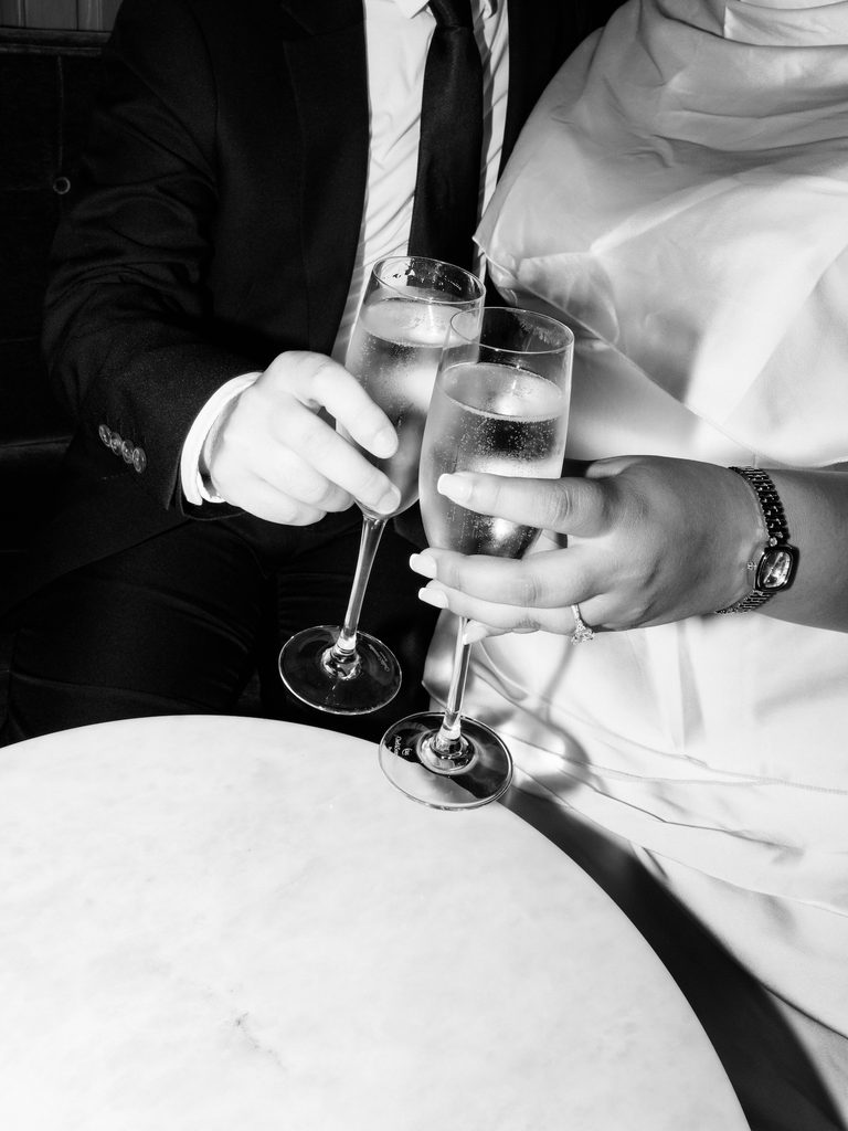 Black-and-white close-up of couple clinking champagne glasses at a small marble table.