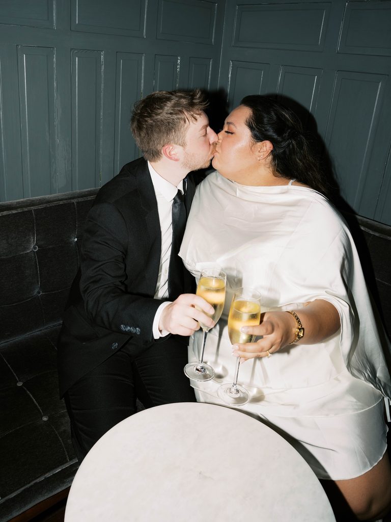 Couple kissing while holding champagne flutes in a dimly lit lounge booth.