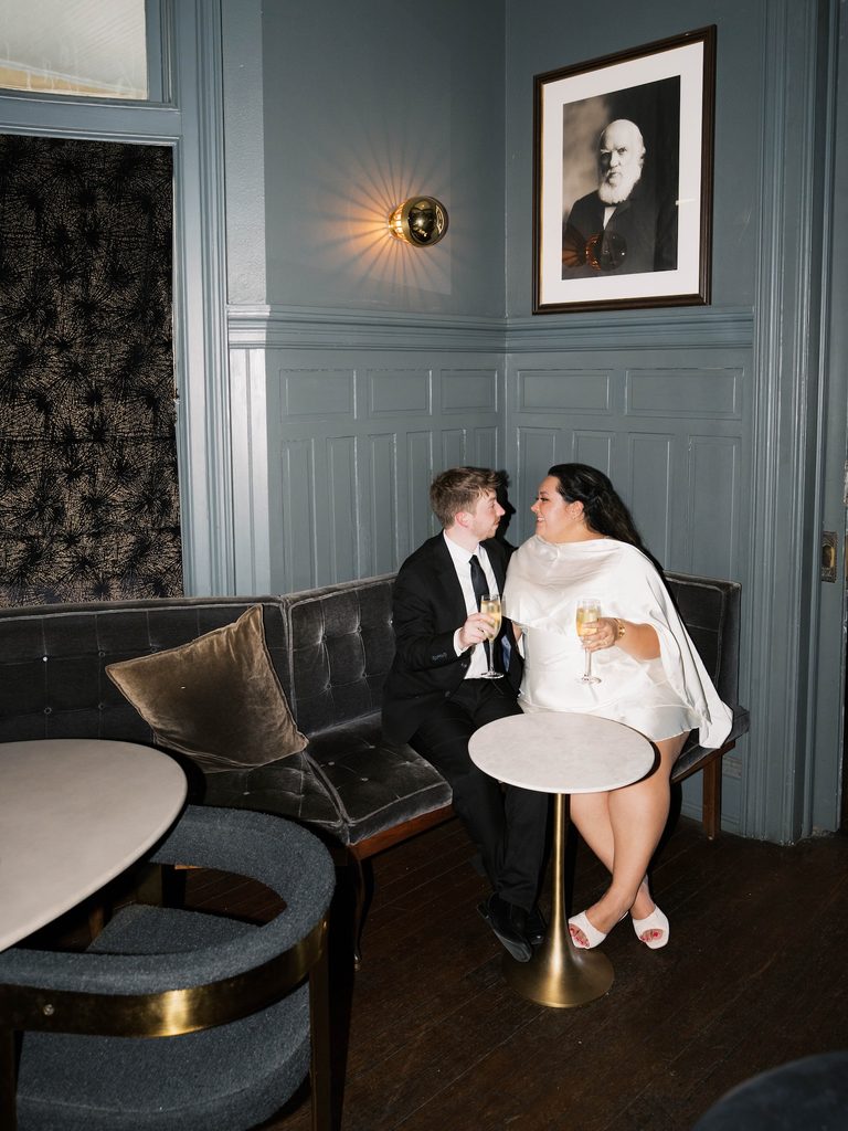 Couple toasting champagne glasses on a velvet lounge sofa, vintage portrait framed on the wall behind them.