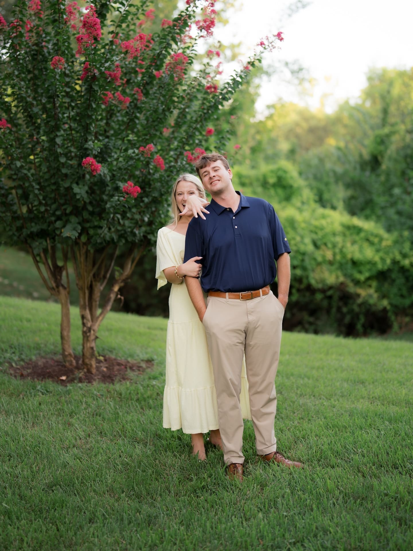 Newly engaged couple laughing and showing off her ring under a blooming crepe myrtle after a surprise proposal at Brenners on the Bayou in Houston
