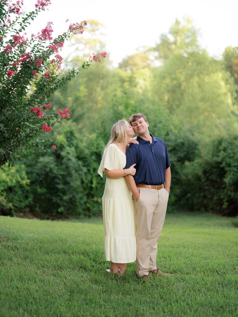 Bride-to-be giving her fiancé a sweet kiss on the cheek while he grins at the camera on the lawn