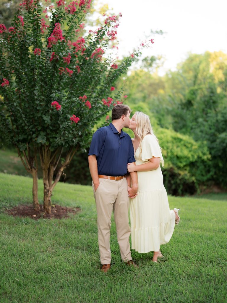 Sweet kiss between a bride-to-be and her fiancé on the lawn, with her heel popped up and pink crepe myrtle blooms behind them