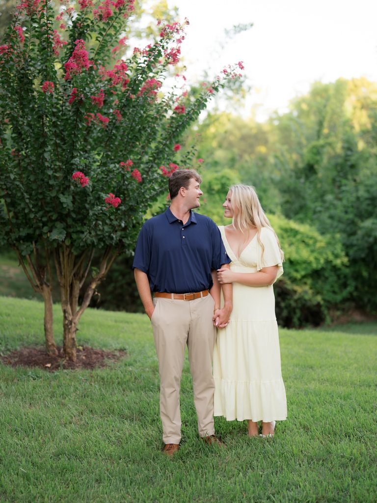 Newly engaged couple holding hands and smiling at each other under a blooming crepe myrtle at their Brenners on the Bayou proposal in Houston