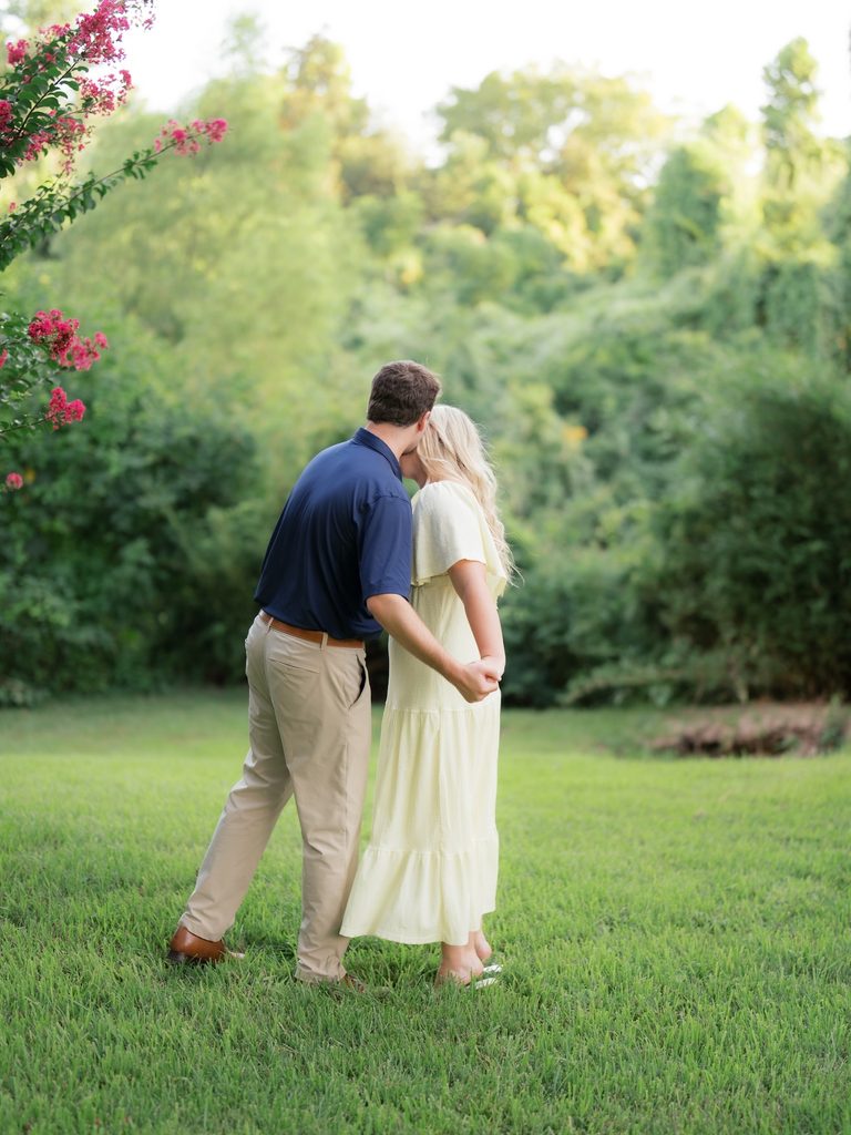 Couple sharing a sweet kiss on the lawn with their backs to the camera, surrounded by greenery and pink crepe myrtle blooms at golden hour