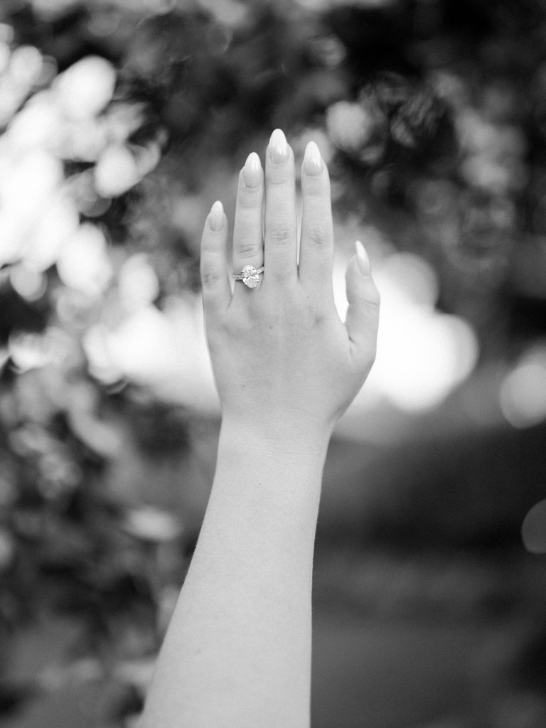 Black and white detail photo of an oval diamond engagement ring on a raised hand with soft, glowing bokeh in the background