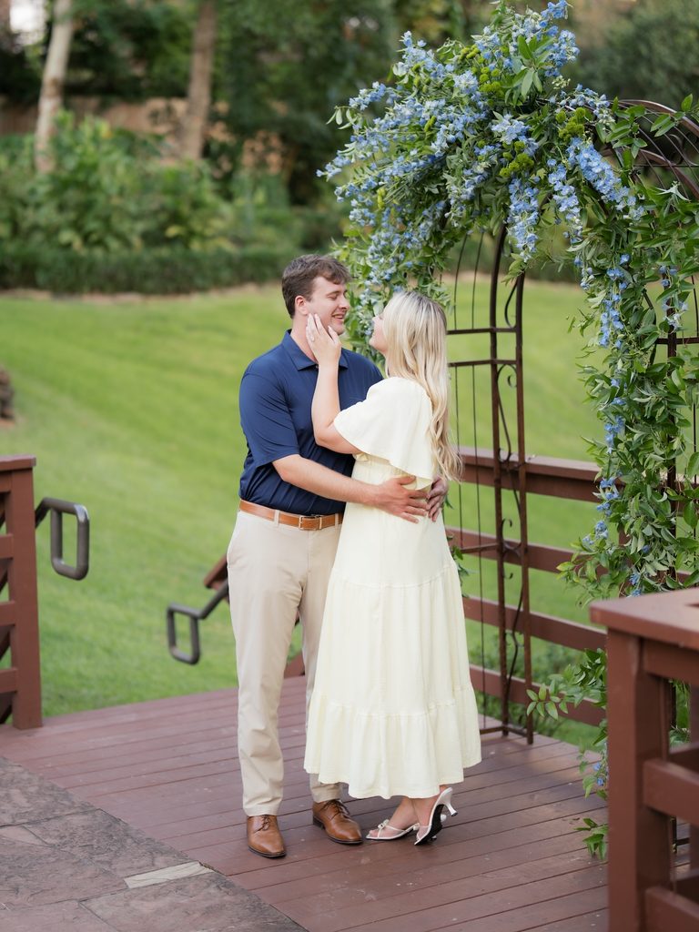 Newly engaged couple in a loving embrace beneath a blue and green floral arch at a Brenners on the Bayou proposal in Houston