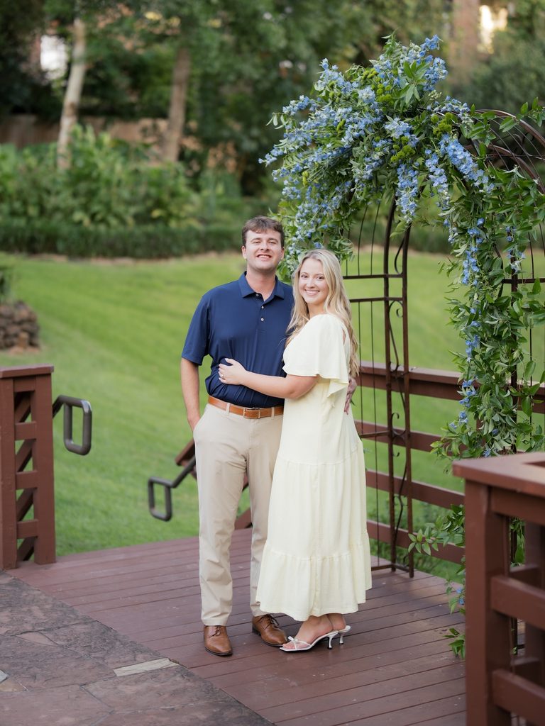 Smiling engaged couple standing together on a wooden deck in front of a blue delphinium floral arch