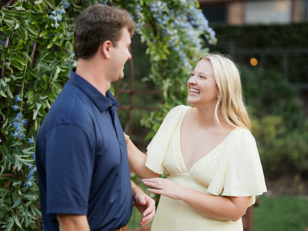 Bride-to-be laughing and admiring her new engagement ring next to her fiancé and a blue floral arch, captured at Brenners on the Bayou