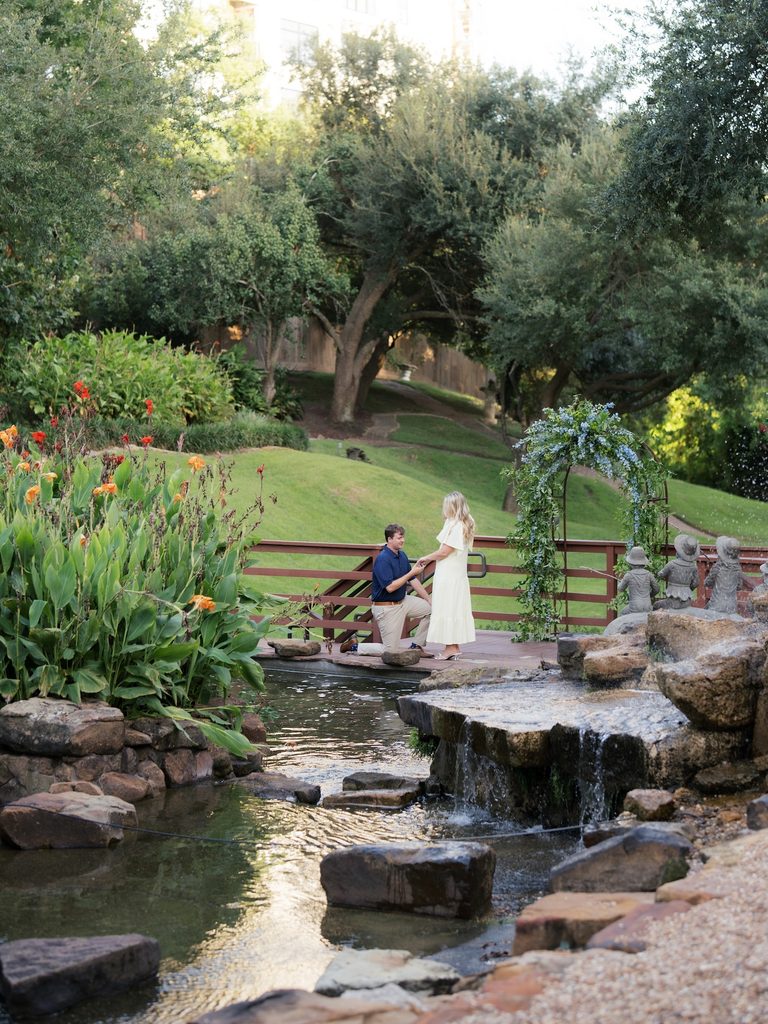 Groom kneeling on a wooden bridge with a blue floral arch, waterfall, and lush garden behind him during a proposal at Brenners on the Bayou