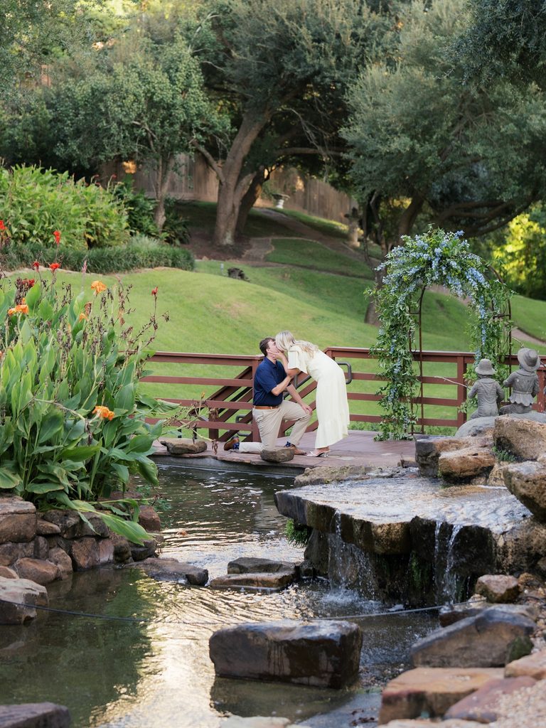 Wide shot of a surprise proposal at Brenners on the Bayou with the couple kissing on a wooden bridge beside a waterfall and floral arch