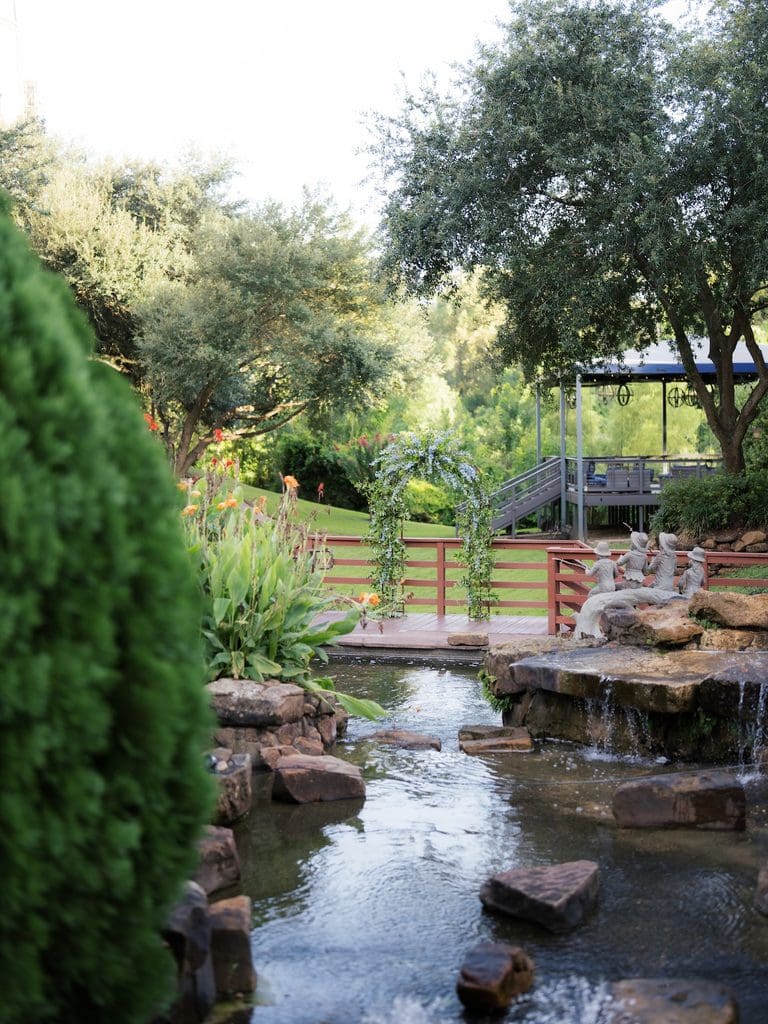 Peaceful view of a proposal setup with a blue floral arch on a wooden bridge, stream, waterfall, and garden statues at golden hour