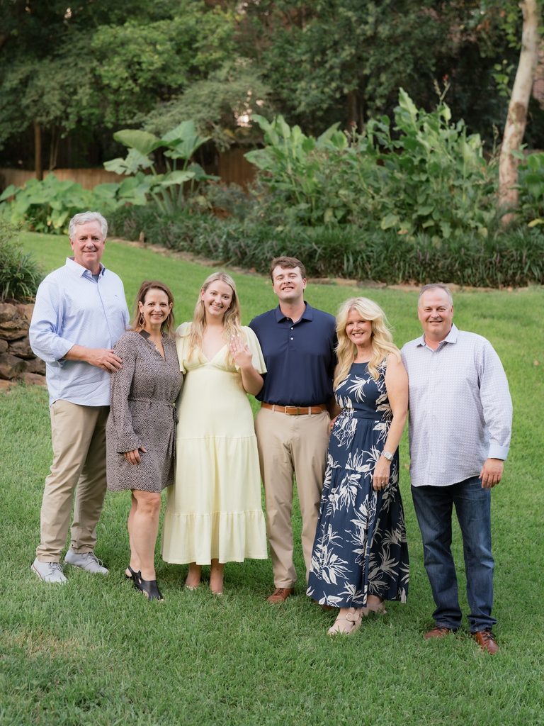 Newly engaged couple showing off the engagement ring while posing with both sets of parents on the lawn at golden hour