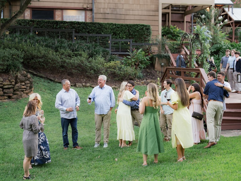 Family and friends gathered on the garden lawn with wine glasses, hugging and celebrating the engagement near the floral arch