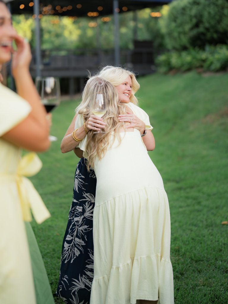 Bride-to-be hugging a guest in a navy floral dress while friends laugh nearby during the engagement celebration