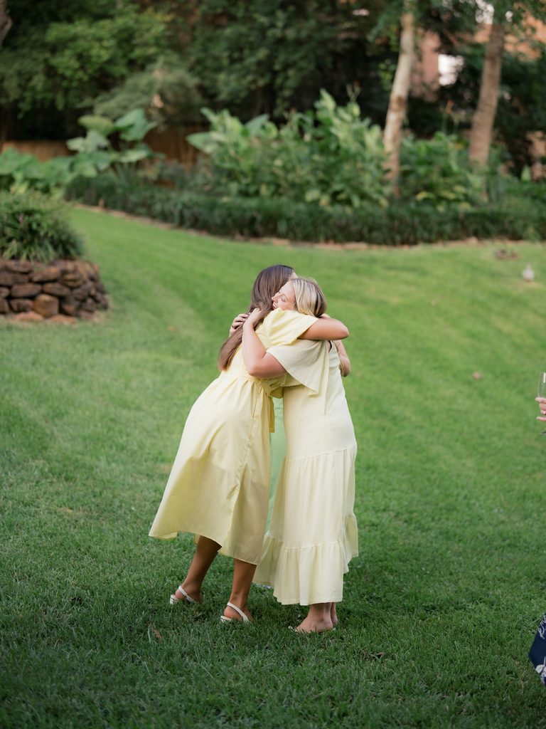 Bride-to-be in a yellow dress sharing a happy hug with a loved one on the lawn after a surprise proposal at Brenners on the Bayou