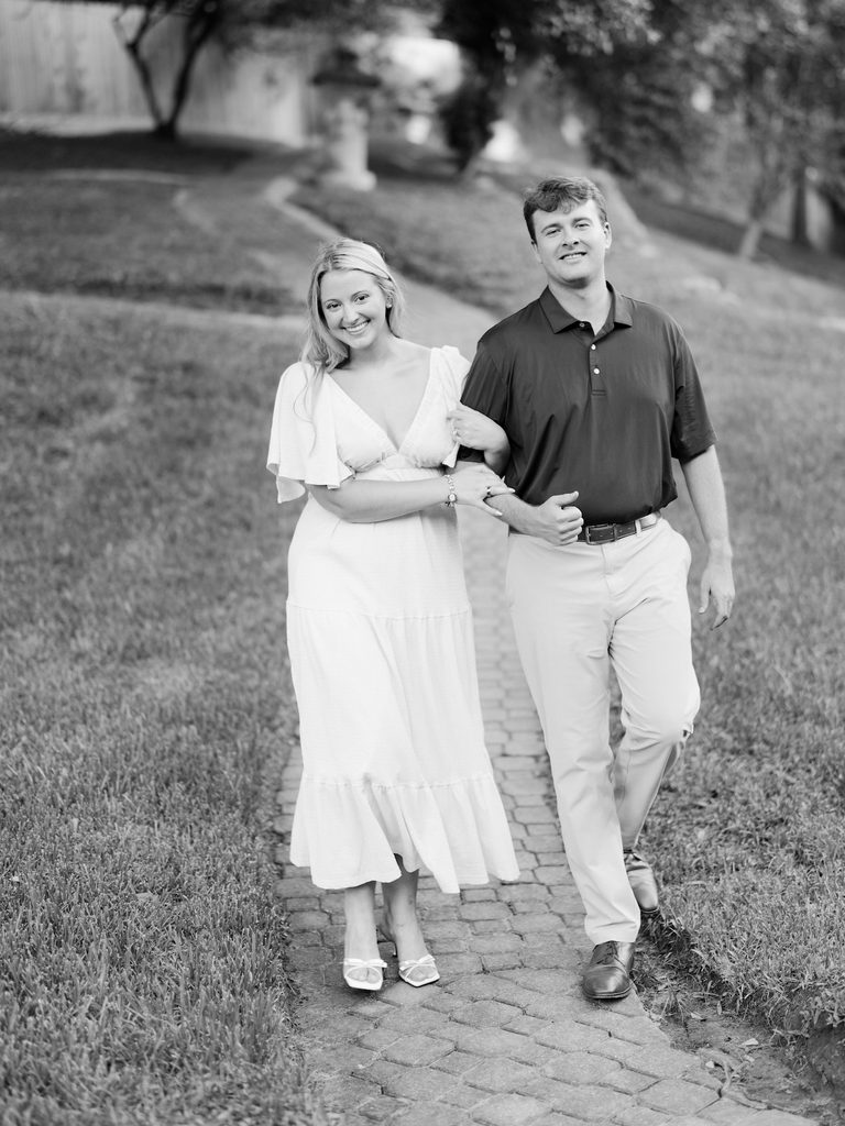 Black and white film-style portrait of an engaged couple walking arm in arm down a brick garden path during their Houston proposal session