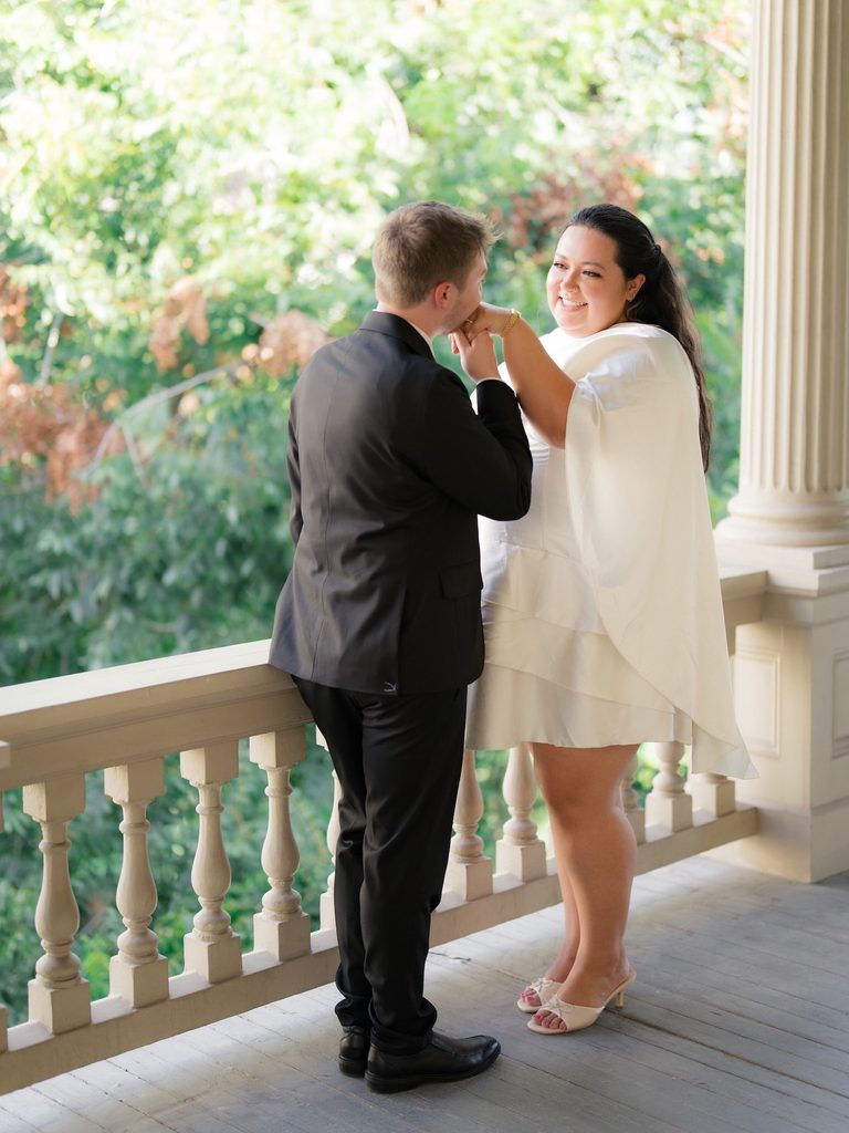 Groom kissing bride's hand on a sunlit balcony framed by tall white columns and greenery.