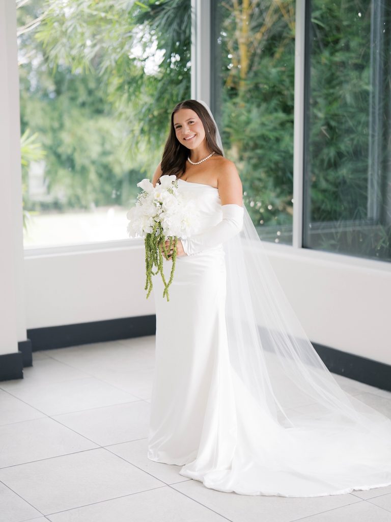 Bridal photos of a smiling bride in a bright sunroom with floor-to-ceiling windows, long veil flowing behind her on white tile