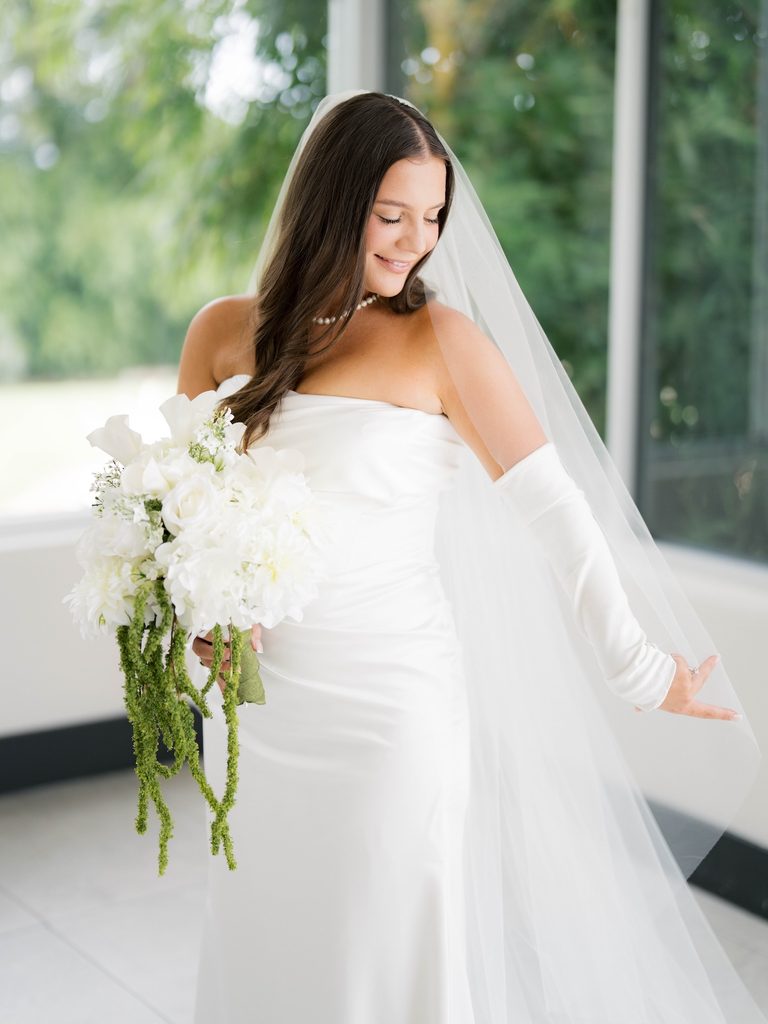 Bridal photos of a bride in a strapless satin gown and long veil, smiling down at her white bouquet with trailing green amaranthus