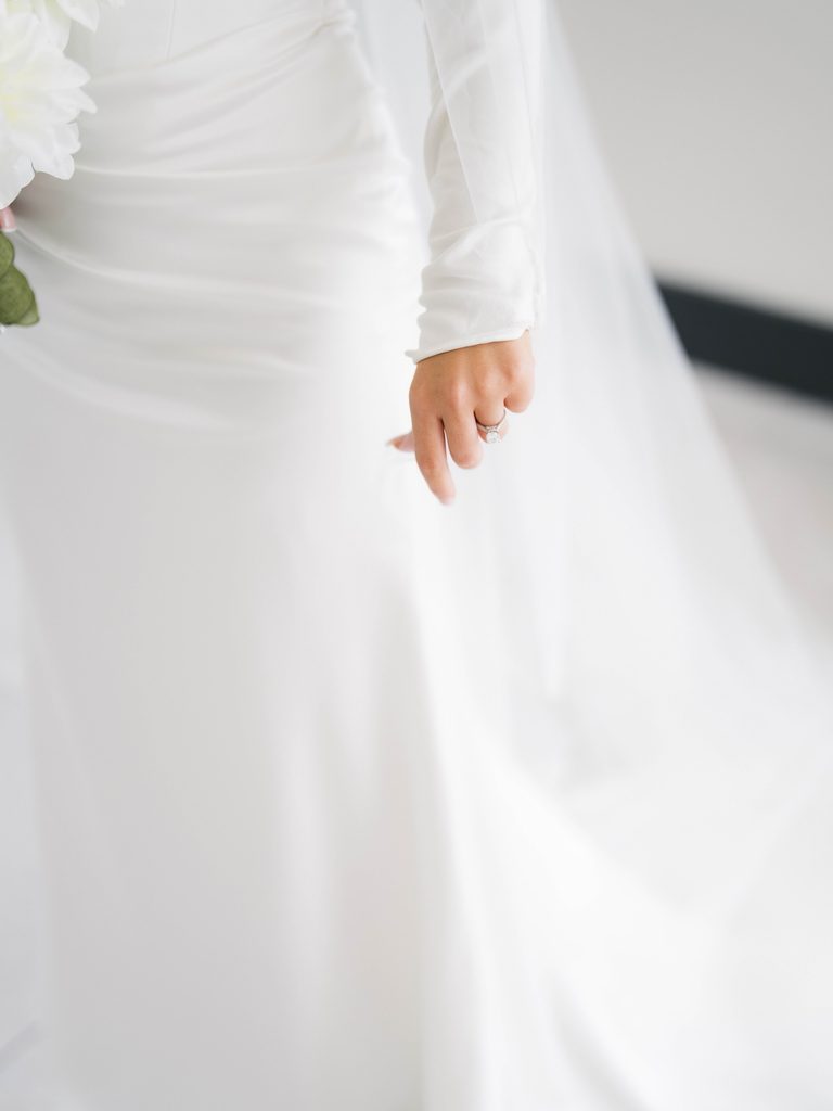 Close-up of a bride’s hand with her engagement ring, resting against a satin gown sleeve and flowing cathedral veil