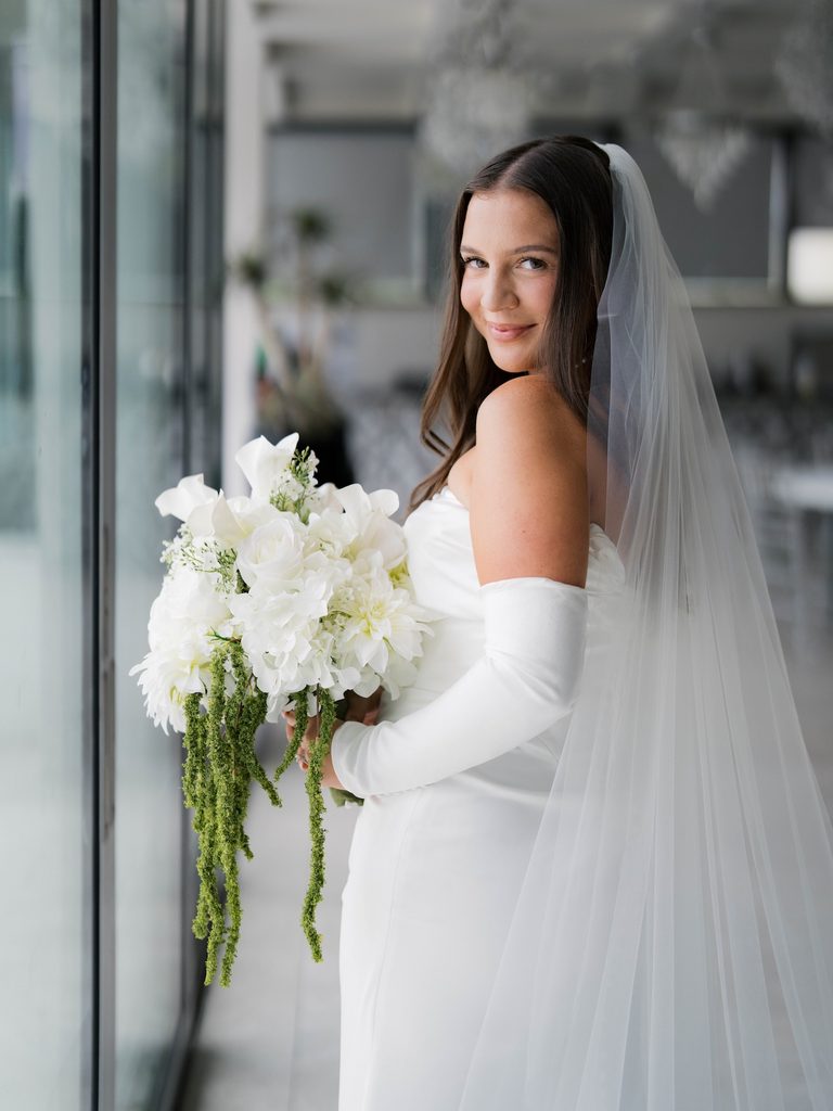 Bride looking back over her shoulder with a soft smile, holding a white bouquet beside a glass wall, veil draped down her back