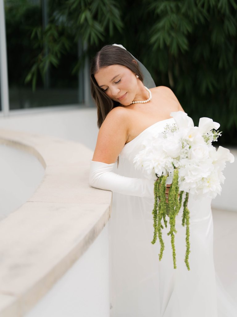 Bride resting her head on her shoulder with her eyes closed, leaning on a curved white ledge with a hanging green bouquet