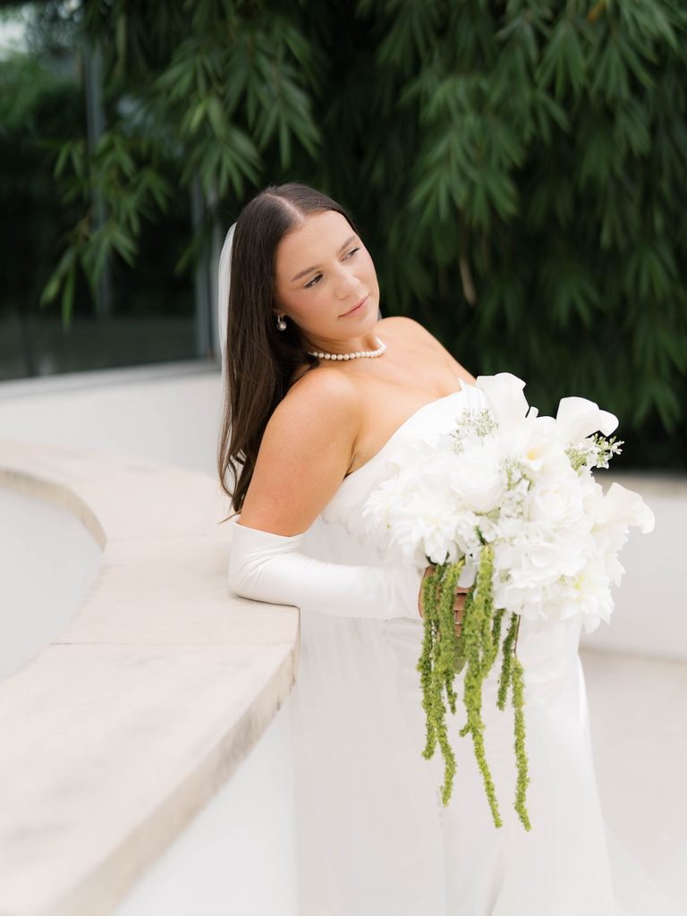 Bridal photos of a bride in pearls leaning on a curved stone ledge with a white calla lily bouquet, bamboo behind her