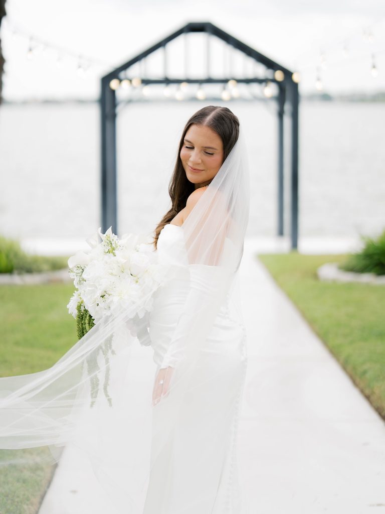 Bridal photos of a bride looking down with her veil blowing, standing on a white path with a dark arbor and waterfront behind her
