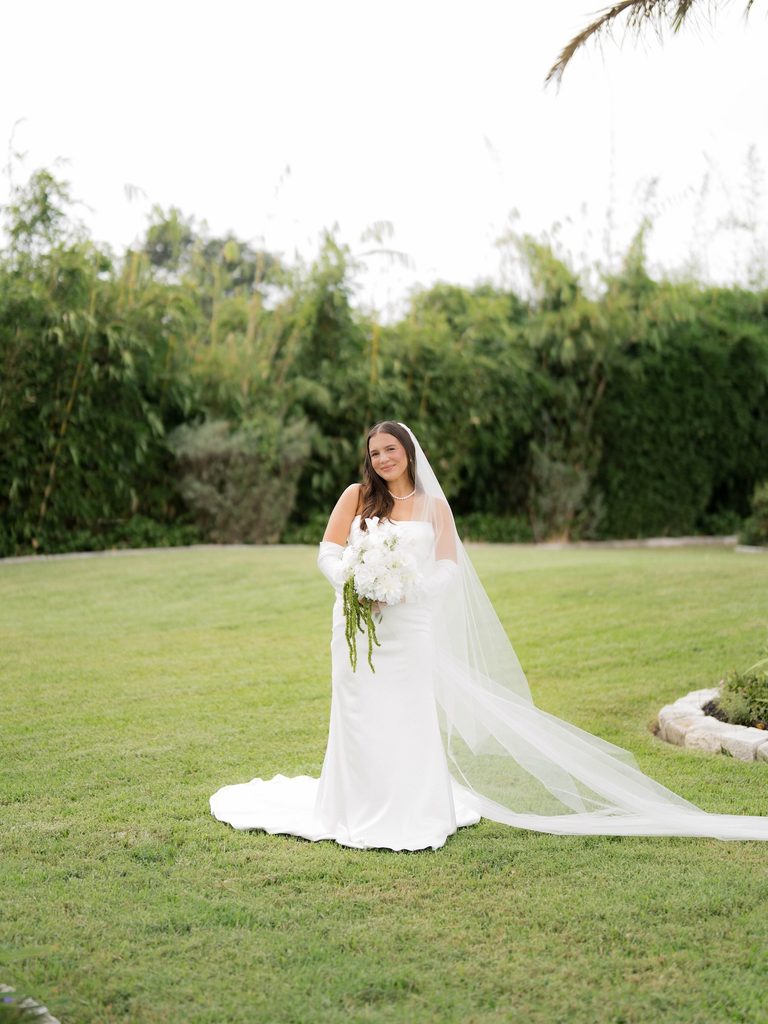 Bride smiling on a green lawn with her head tilted, holding a white bouquet with trailing green amaranthus, long veil flowing beside her