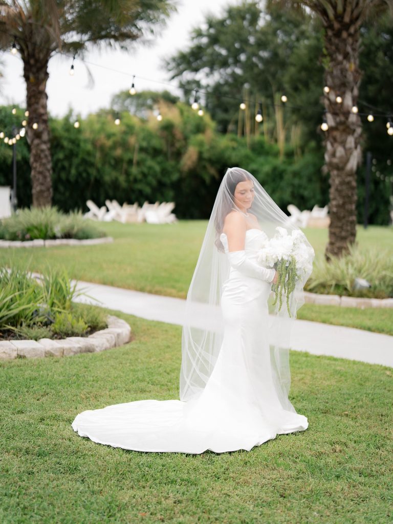 Bride on a green lawn with her veil pulled over her face, satin train spread out, palm trees and string lights behind