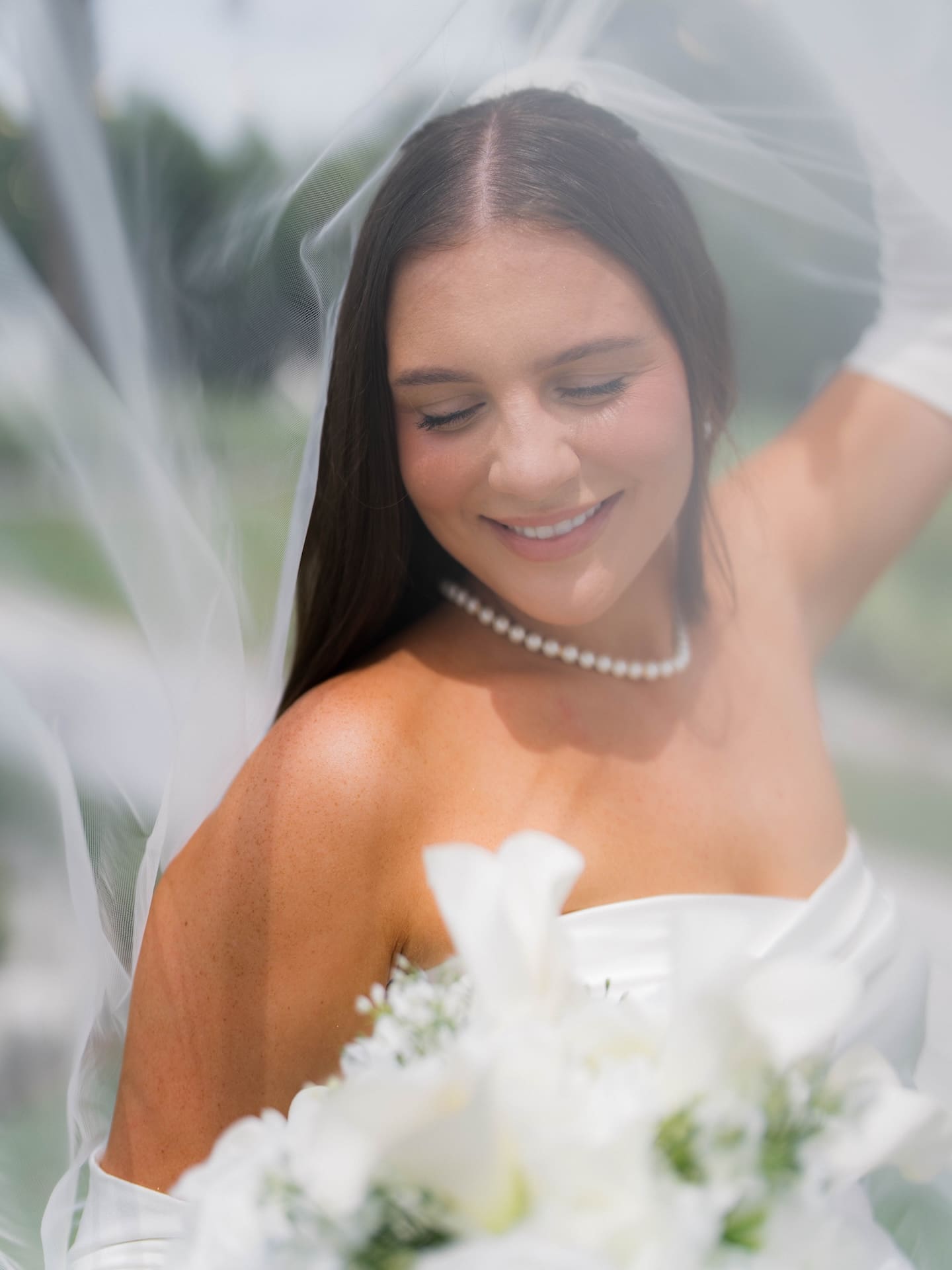 Bride smiling down towards her flowers under a sheer veil, pearl necklace on, soft white bouquet blurred in the foreground