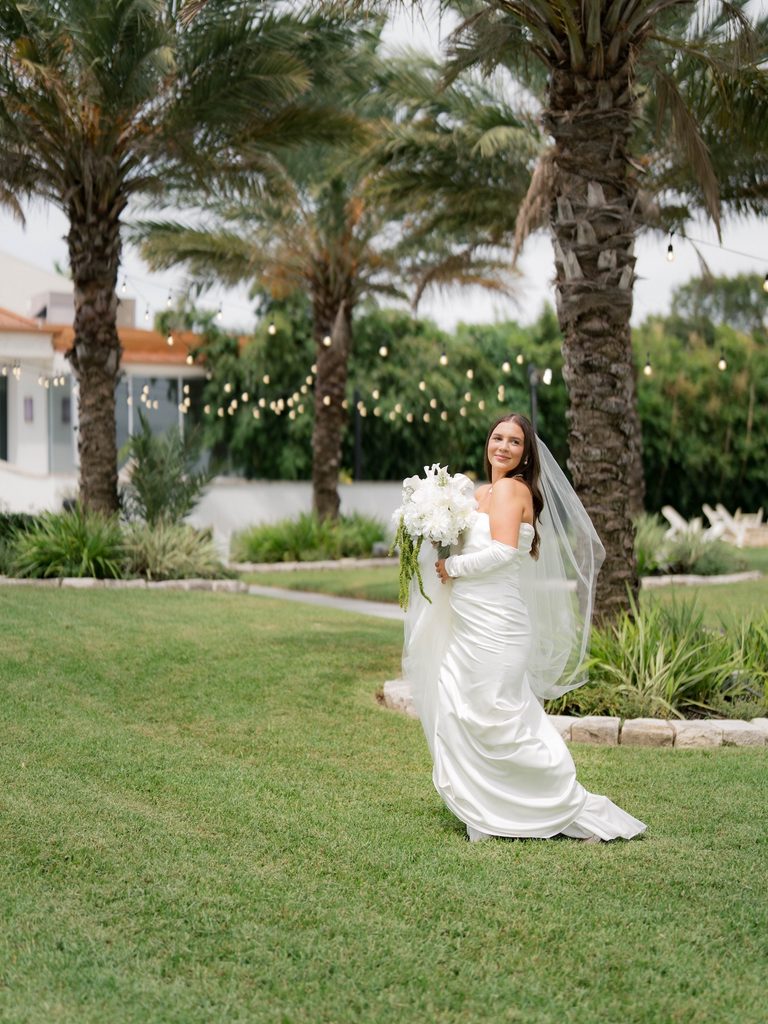 Bridal photos of a bride glancing over her shoulder on a lawn lined with palm trees and string lights, satin gown and veil moving in the breeze