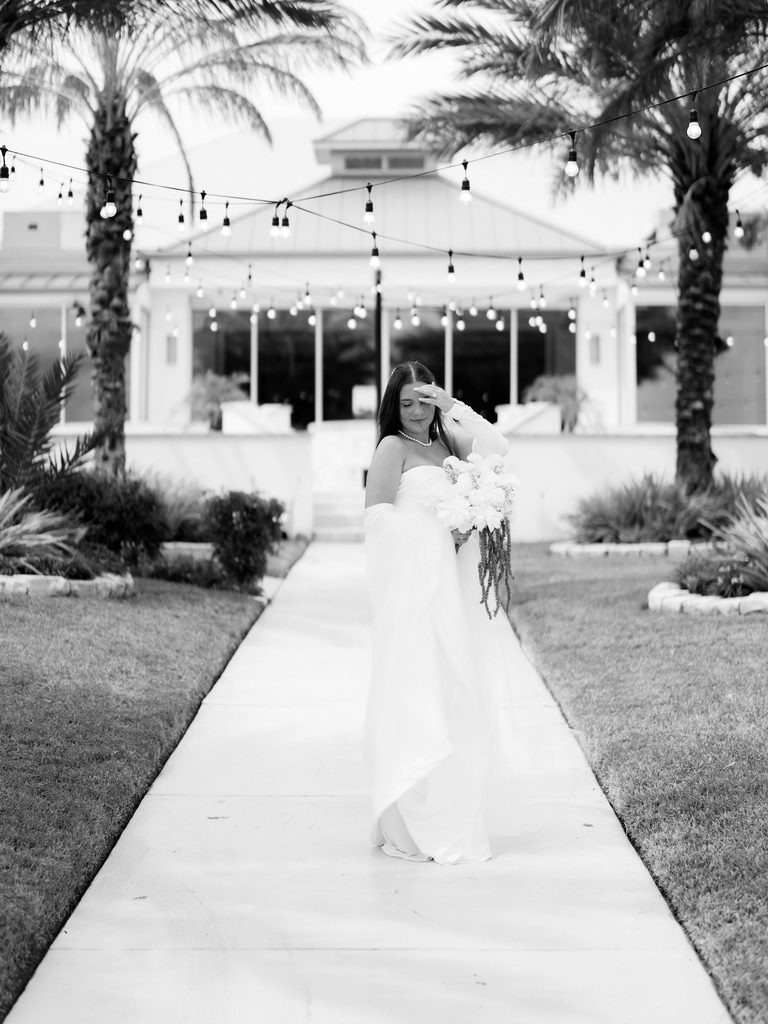 Black and white photo of a bride on a walkway under string lights, hand at her forehead, palm trees and a white building behind