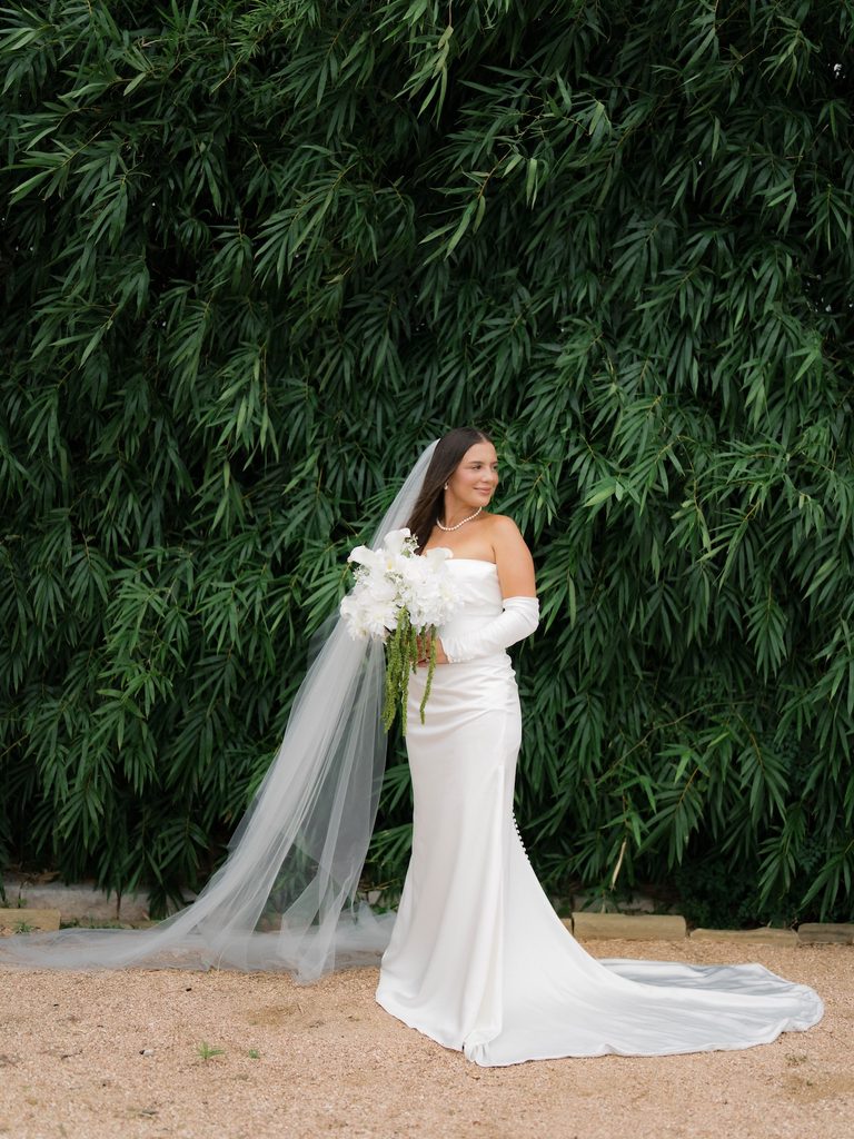 Full-length bride in a satin gown with a long train and veil, standing in front of a lush bamboo wall