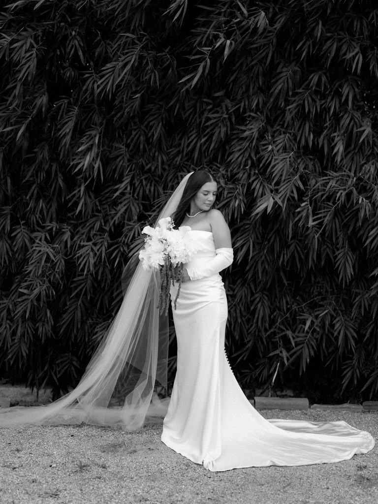 Black and white bridal photos of a bride looking down over her shoulder in front of a dark bamboo wall, long veil and satin train spread across the gravel