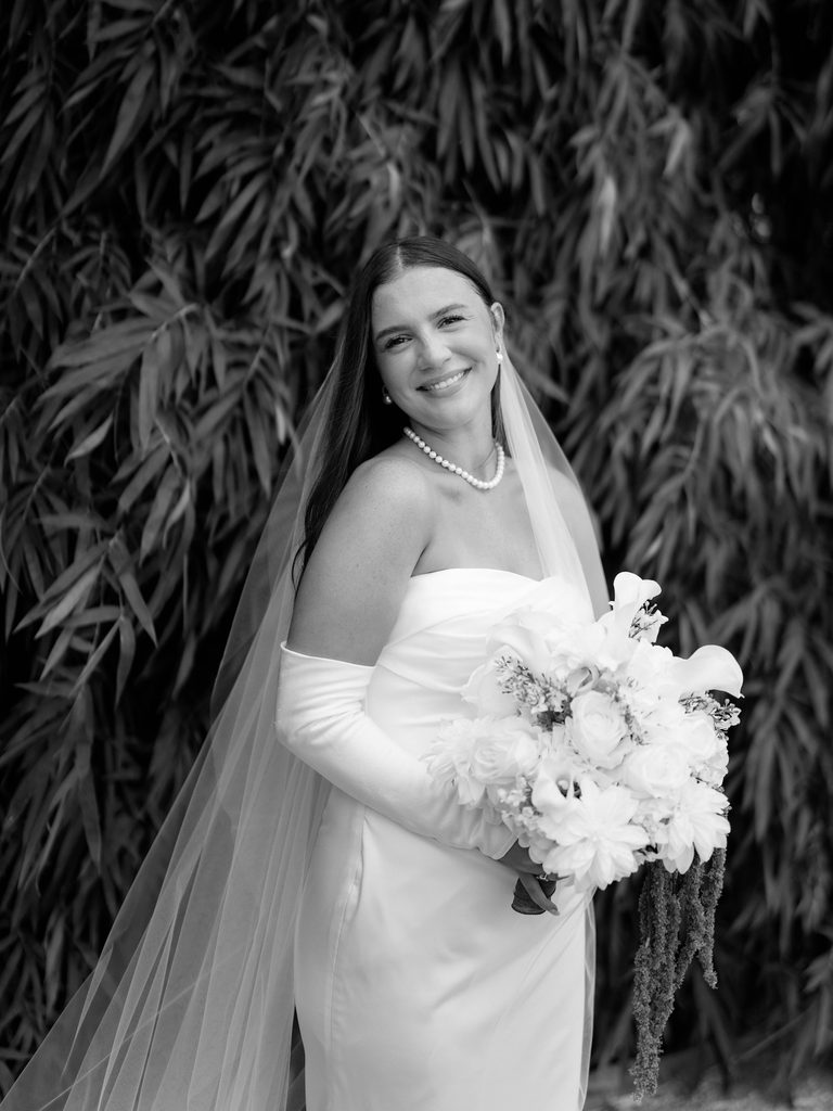 Black and white bridal photos of a smiling bride with a pearl necklace, veil, and white bouquet in front of bamboo leaves