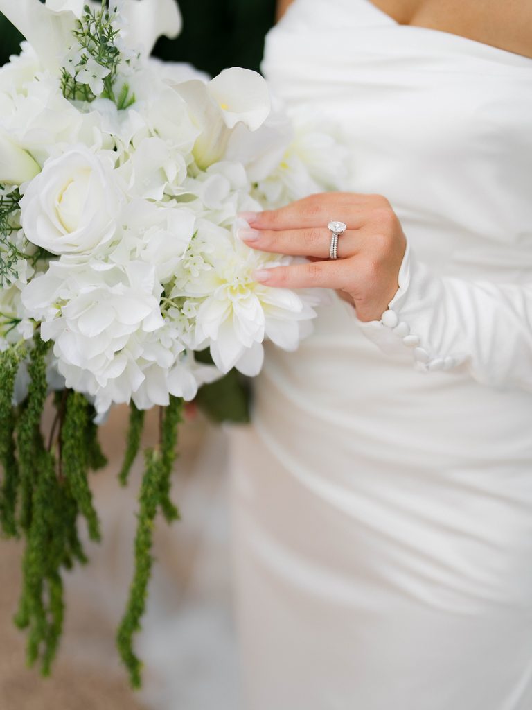 Bridal photos detail of a bride’s oval engagement ring and wedding band resting on white roses, dahlias, and calla lilies