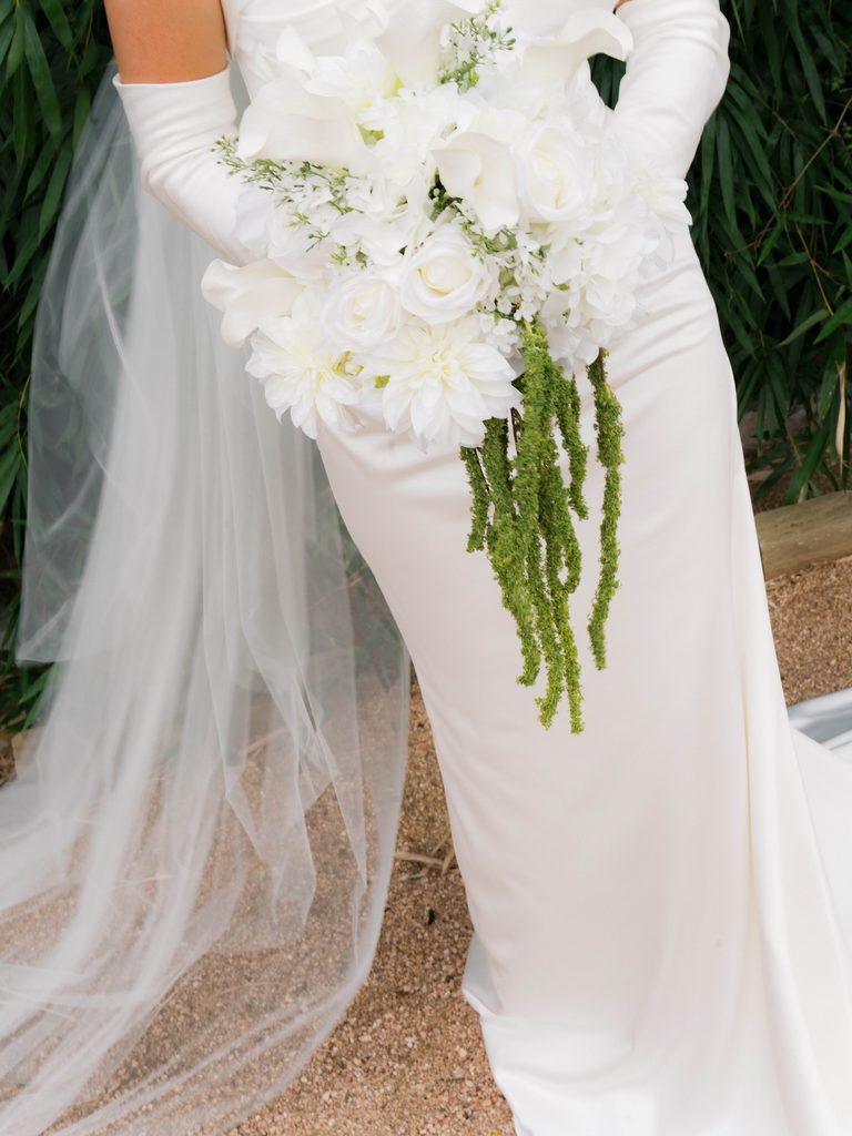 Detail of a bride’s hanging bouquet and button-back satin gown with a sheer veil on a gravel path