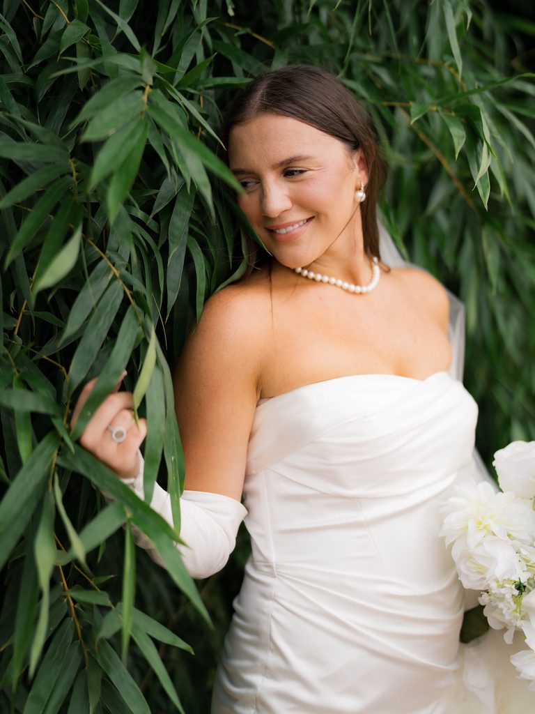 Bridal photos of a bride leaning into green bamboo leaves and glancing over her shoulder, wearing opera-length satin gloves