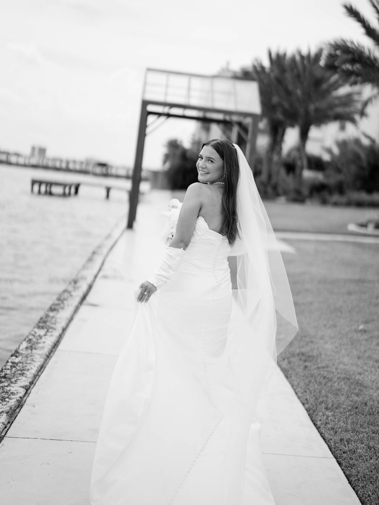Bridal photos of a bride in black and white walking along the waterfront, looking back and smiling, holding her veil, buttons down her gown
